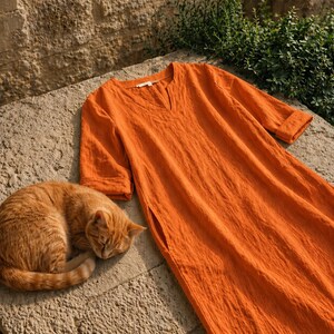 Puede incluir: Un vestido de lino naranja con cuello en V y mangas hasta el codo descansa sobre una superficie de piedra. Un gato atigrado naranja durmiendo est&aacute; acurrucado junto al vestido. El vestido tiene un corte holgado y bolsillos laterales.