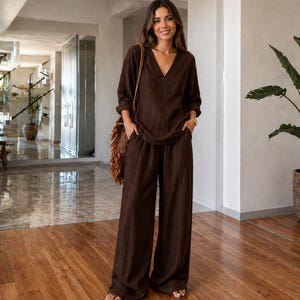 May include: A brown linen outfit consisting of a V-neck top and wide-leg pants. The outfit is worn with a woven shoulder bag and sandals. The outfit is a solid color and appears to be made of a lightweight fabric.