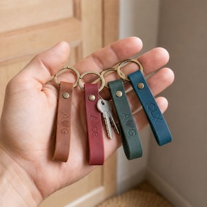 May include: Four leather keychains in tan, maroon, dark green, and blue, each featuring a metal ring and a brass rivet. The tan keychain is embossed with A, a heart, and G. The maroon one says Alice. The green one has A, a heart, and G. The blue one says CLX.