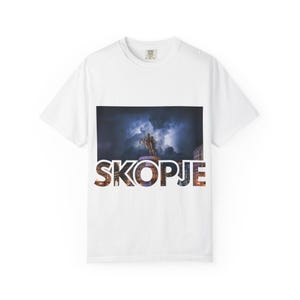 Könnte beinhalten: Weißes T-Shirt mit einer Grafik einer Statue und dem Wort "SKOPJE" in bunten Buchstaben. Das Bild auf dem Shirt zeigt eine Statue einer Person auf einem Pferd unter einem dunklen, bewölkten Himmel. Das T-Shirt ist aus einem weichen Material.