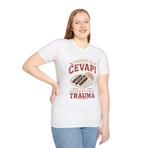 May include: White t-shirt with a graphic design featuring the text "Powered by Ćevapi and Generational Trauma." The design includes an illustration of ćevapi, a Balkan dish, on a plate. The shirt is short-sleeved.