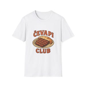 Peut inclure: Un t-shirt blanc à manches courtes avec une illustration graphique de saucisses grillées servies sur une assiette avec du pain plat. Le texte "ČEVAPI CLUB" est imprimé en lettres capitales orange au-dessus et en dessous de l'illustration.