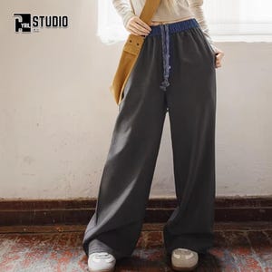 Könnte beinhalten: Weit geschnittene Hose in Dunkelanthrazit mit blauem elastischem Bund und passendem blauem Kordelzug. Die Hose hat eine lockere Passform und wird mit einem langärmeligen weißen Oberteil und einer beigen Umhängetasche kombiniert. Cyrill Studio Logo sichtbar.