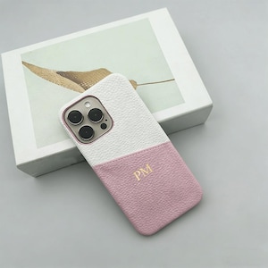 Puede incluir: Funda de teléfono de dos tonos en cuero blanco y rosa. La mitad superior es blanca y la mitad inferior es rosa. Las iniciales "PM" están grabadas en oro en la sección rosa. La funda está sobre una caja blanca.