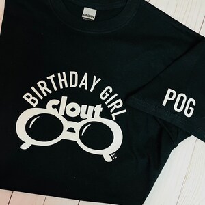 May include: Black t-shirt with white text and graphics. The shirt features the words "BIRTHDAY GIRL clout" in a circular design, with a pair of glasses in the center. The number "12" is below the glasses. The sleeve has the word "POG".