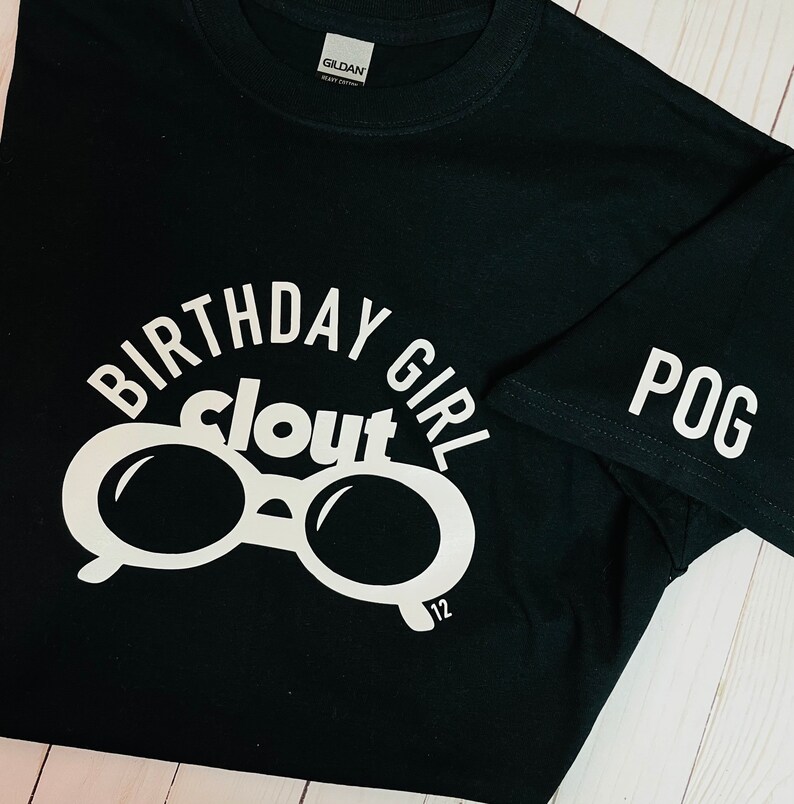 May include: Black t-shirt with white text and graphics. The shirt features the words "BIRTHDAY GIRL clout" in a circular design, with a pair of glasses in the center. The number "12" is below the glasses. The sleeve has the word "POG".
