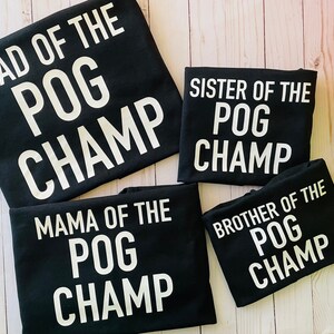 May include: Four black t-shirts with white text that reads "DAD OF THE POG CHAMP", "SISTER OF THE POG CHAMP", "MAMA OF THE POG CHAMP", and "BROTHER OF THE POG CHAMP". The shirts are arranged on a light wood surface.