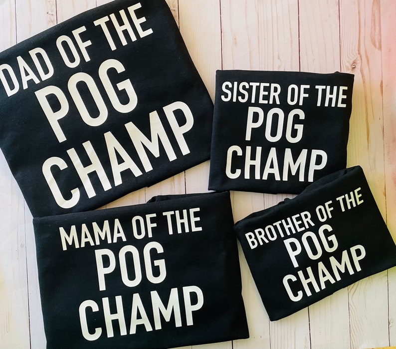May include: Four black t-shirts with white text that reads "DAD OF THE POG CHAMP", "SISTER OF THE POG CHAMP", "MAMA OF THE POG CHAMP", and "BROTHER OF THE POG CHAMP". The shirts are arranged on a light wood surface.