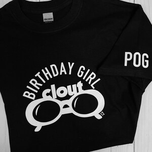 May include: Black t-shirt with white text and graphics. The shirt features the words "BIRTHDAY GIRL clout" above a pair of sunglasses. The sleeve has the word "POG" printed on it. The number "12" is printed on the sunglasses.