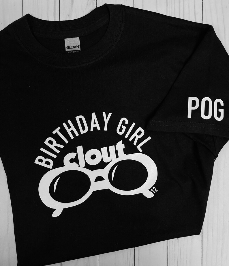 May include: Black t-shirt with white text and graphics. The shirt features the words "BIRTHDAY GIRL clout" above a pair of sunglasses. The sleeve has the word "POG" printed on it. The number "12" is printed on the sunglasses.