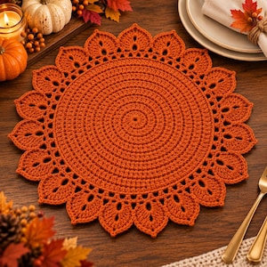 Può includere: Una tovaglietta rotonda all'uncinetto color arancio bruciato, caratterizzata da un centro denso a spirale e un bordo decorativo composto da forme ripetitive a petalo. Il tessuto è realizzato con un motivo a punti uniforme.