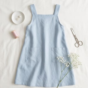 May include: A light blue sleeveless apron dress featuring a square neckline, wide shoulder straps, and two large patch pockets on the front. The garment is displayed alongside a small white dish with two buttons, a spool of pink thread, and scissors.