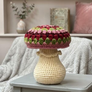 Peut inclure: Lampe champignon en crochet avec un pied crème et un chapeau coloré. Le chapeau présente un motif patchwork dans les tons de rose, de rouge et de vert. La lampe est posée sur une table grise.