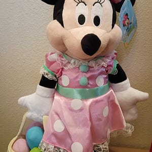 May include: A Minnie Mouse plush doll wearing a pink dress with white polka dots, a matching bow, and a basket of Easter eggs. The doll has black ears, a pink face, and white gloves. The dress has lace trim and a mint green sash.