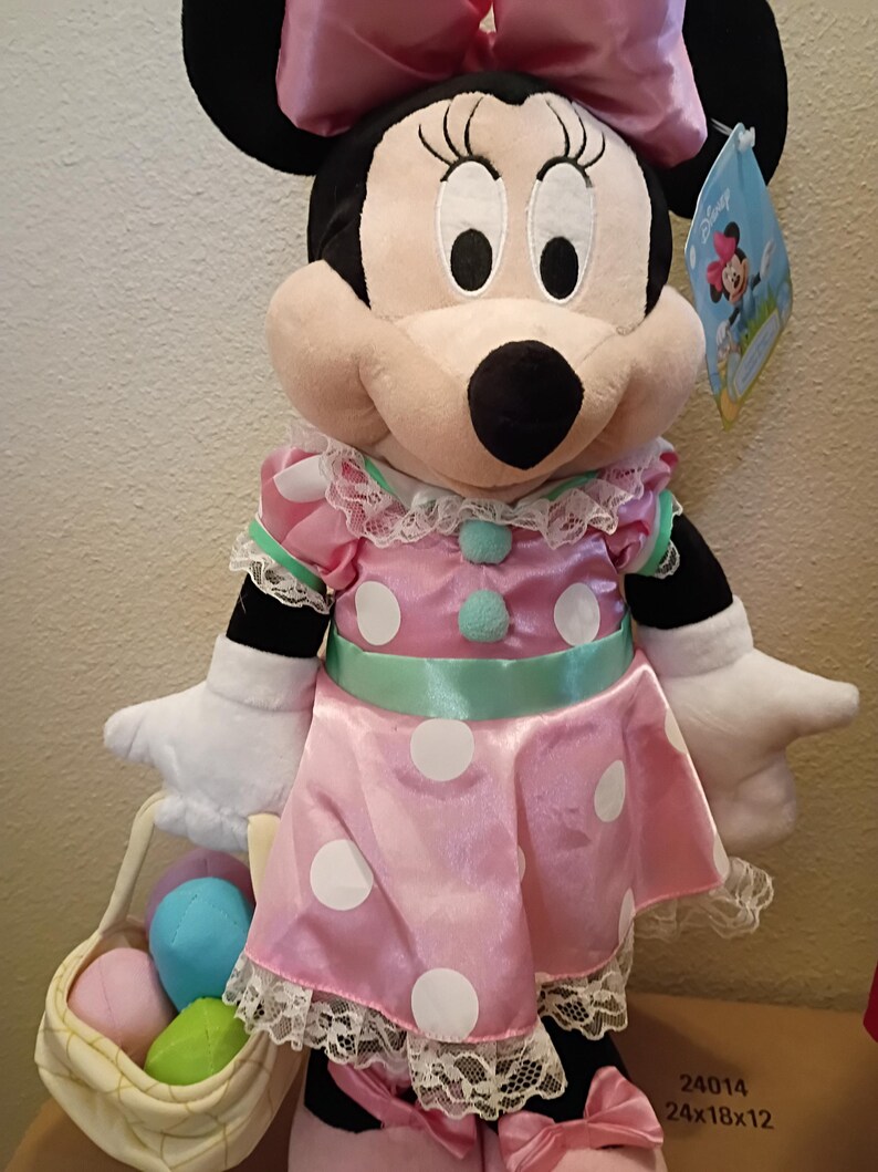 May include: A Minnie Mouse plush doll wearing a pink dress with white polka dots, a matching bow, and a basket of Easter eggs. The doll has black ears, a pink face, and white gloves. The dress has lace trim and a mint green sash.