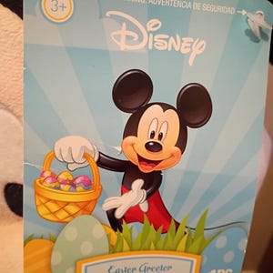 May include: A Disney Easter Greeter tag featuring Mickey Mouse holding a basket of colorful Easter eggs. The tag has a light blue background with the Disney logo and the text "Easter Greeter, Saludaor de Pascua".