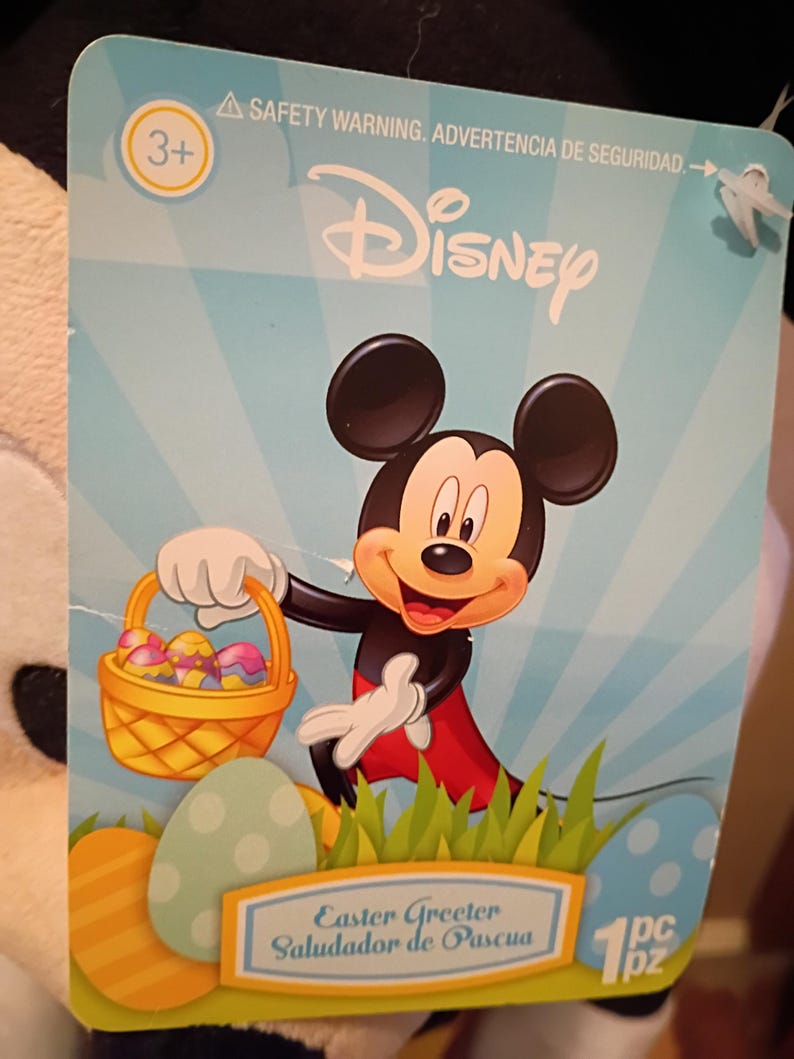 May include: A Disney Easter Greeter tag featuring Mickey Mouse holding a basket of colorful Easter eggs. The tag has a light blue background with the Disney logo and the text "Easter Greeter, Saludaor de Pascua".