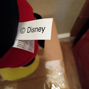 May include: A plush toy with a red body, black head, and yellow feet. A white tag with the "&copy; Disney" logo is attached to the toy. The toy is partially visible, resting on a cardboard surface.