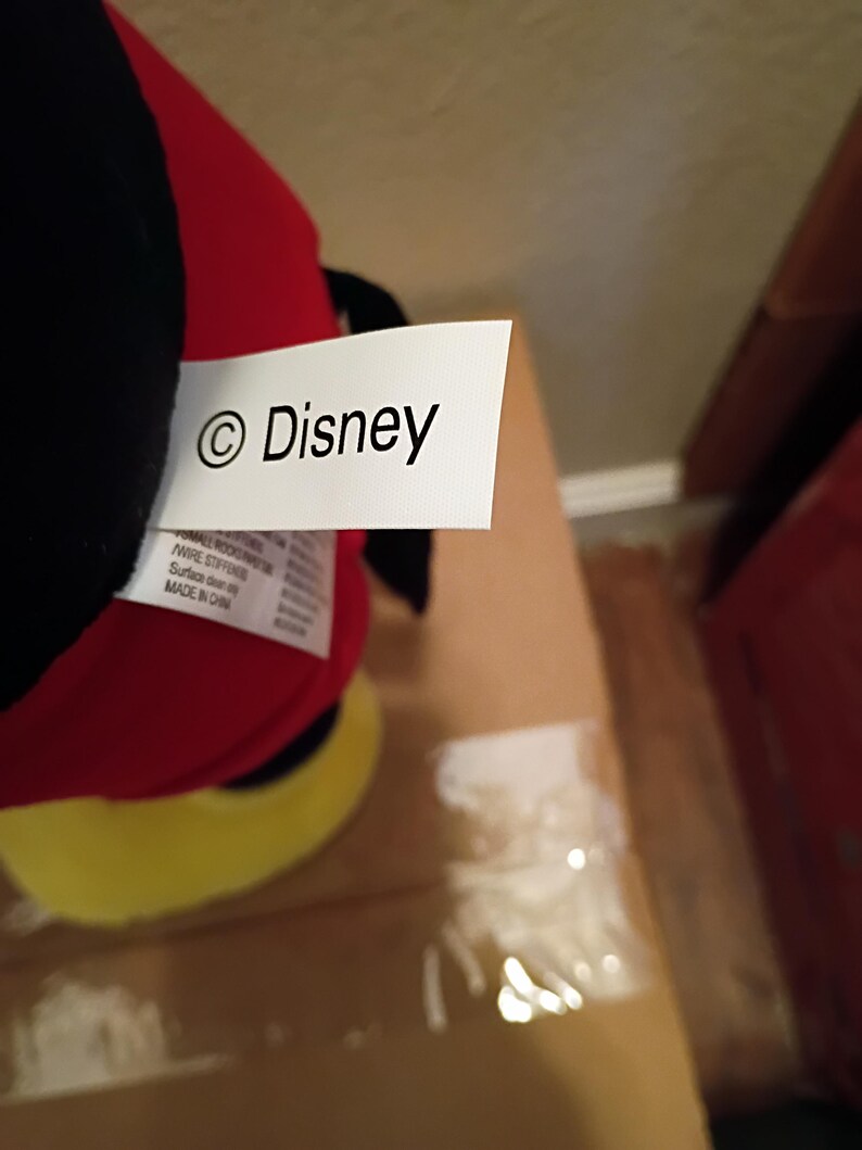May include: A plush toy with a red body, black head, and yellow feet. A white tag with the "&copy; Disney" logo is attached to the toy. The toy is partially visible, resting on a cardboard surface.