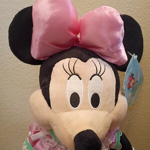 May include: A plush Minnie Mouse doll with a large pink satin bow, black ears, and a pink polka-dot dress. The doll has a white lace collar and a tag attached. The doll is a classic Disney character.