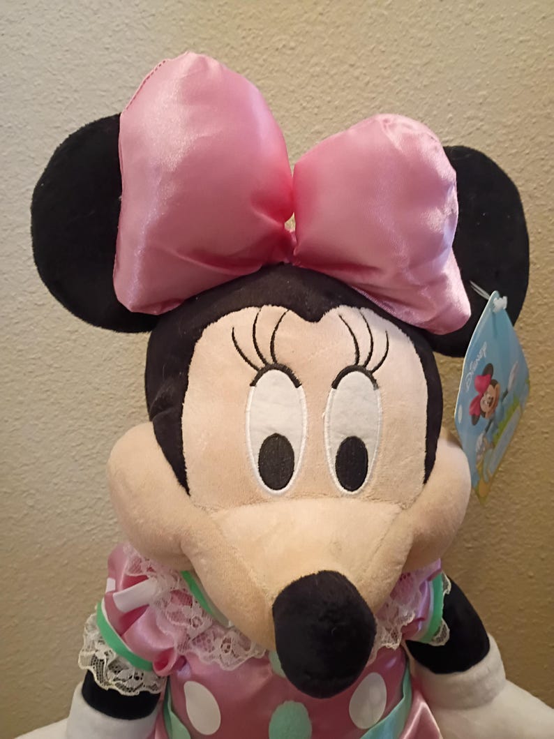 May include: A plush Minnie Mouse doll with a large pink satin bow, black ears, and a pink polka-dot dress. The doll has a white lace collar and a tag attached. The doll is a classic Disney character.