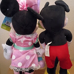 May include: Two plush Disney character dolls, Minnie Mouse and Mickey Mouse, are shown from the back. Minnie wears a pink polka-dot dress with a matching bow. Mickey wears red shorts and yellow shoes. Both have black ears and tails.