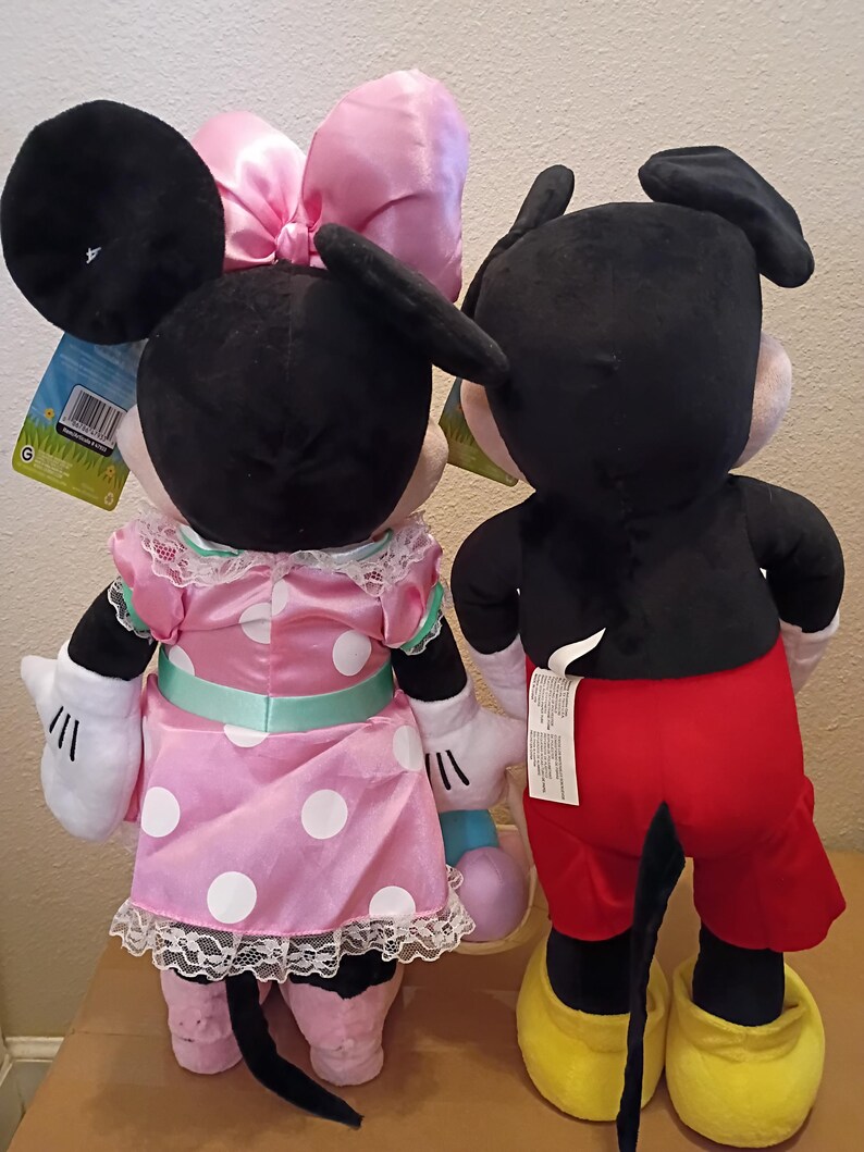 May include: Two plush Disney character dolls, Minnie Mouse and Mickey Mouse, are shown from the back. Minnie wears a pink polka-dot dress with a matching bow. Mickey wears red shorts and yellow shoes. Both have black ears and tails.