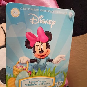 May include: A Disney Easter Greeter tag featuring Minnie Mouse with a pink bow, blue dress, and a basket of Easter eggs. The tag has the Disney logo and the text "Easter Greeter" and "Saludador de Pascua".