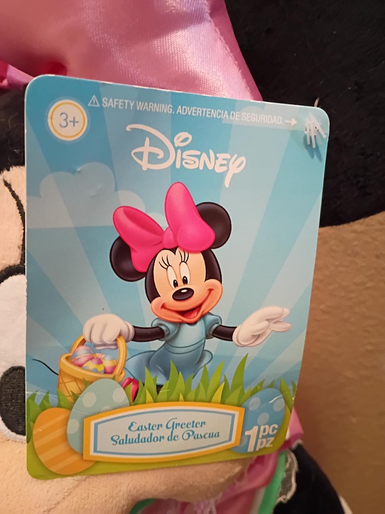 May include: A Disney Easter Greeter tag featuring Minnie Mouse with a pink bow, blue dress, and a basket of Easter eggs. The tag has the Disney logo and the text "Easter Greeter" and "Saludador de Pascua".