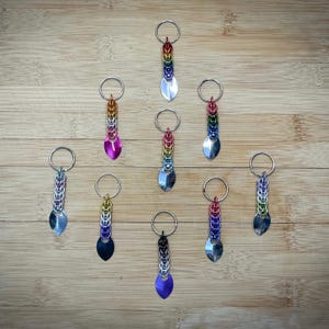 May include: Ten colorful keychains with silver rings and a silver leaf-shaped charm. Each keychain is made of woven metal rings in a rainbow color pattern.