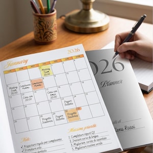 Può includere: Un'agenda 2026 aperta a gennaio, con un layout a calendario e note manoscritte. L'agenda è su una superficie di legno, con una penna in mano e un portapenne.