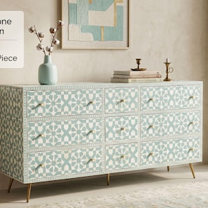 Op de afbeelding: Een rechthoekige ladekast met een geometrisch patroon van lichtblauw en wit beeninlegwerk over negen lades. Het meubel heeft kleine goudkleurige knoppen op elke lade en rust op vier dunne, taps toelopende goudkleurige metalen poten.
