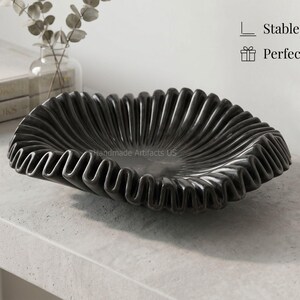 May include: A black decorative bowl with a wide, ruffled, and pleated edge. The bowl features a deep, circular centre and a textured, wavy rim. The text on the image includes Stable & Heavy Base, Perfect for Gifting, and Handcrafted Design.