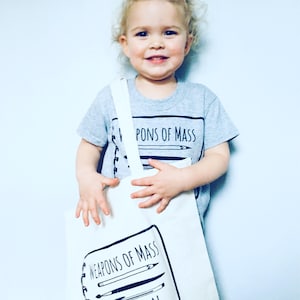 Puede incluir: Un ni&ntilde;o lleva una camiseta gris con el texto "Weapons of Mass Creation" y sostiene una bolsa de tela blanca con el mismo texto y un dibujo de &uacute;tiles de arte.