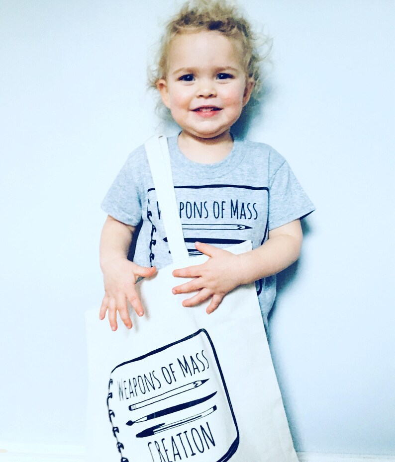 Puede incluir: Un ni&ntilde;o lleva una camiseta gris con el texto "Weapons of Mass Creation" y sostiene una bolsa de tela blanca con el mismo texto y un dibujo de &uacute;tiles de arte.