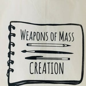 Puede incluir: Una bolsa de lona blanca con una ilustraci&oacute;n en blanco y negro de un cuaderno con el texto "Weapons of Mass Creation" y un l&aacute;piz, un bol&iacute;grafo y un pincel.