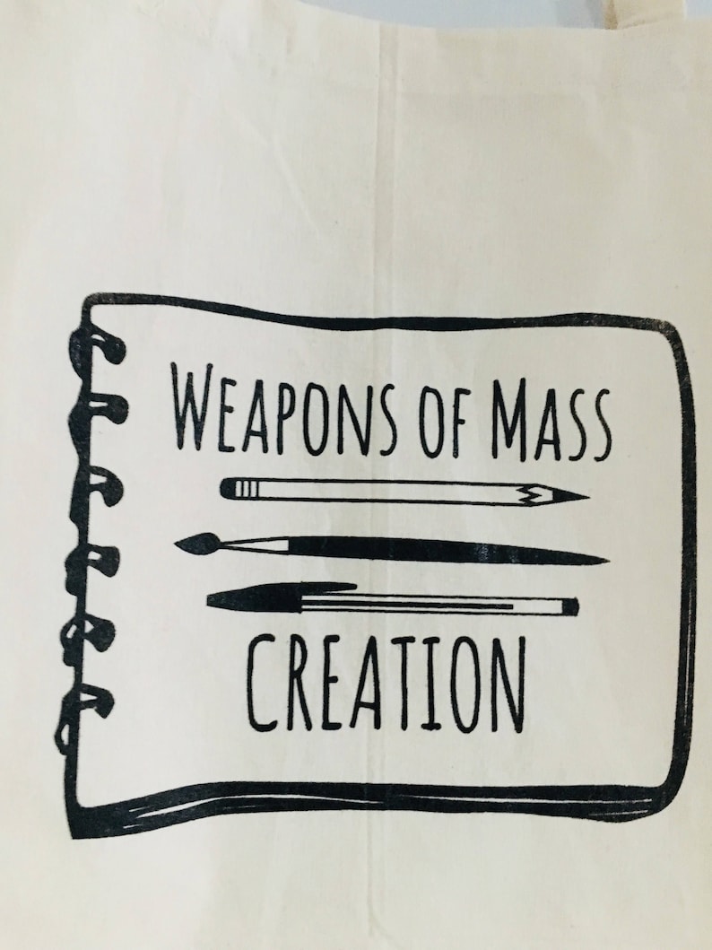 Puede incluir: Una bolsa de lona blanca con una ilustraci&oacute;n en blanco y negro de un cuaderno con el texto "Weapons of Mass Creation" y un l&aacute;piz, un bol&iacute;grafo y un pincel.