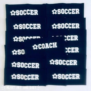 以下が含まれることがあります： 白いサッカーボールと文字が書かれた黒い布製のリストバンドの山。リストバンドの文字は「SOCCER」と「COACH」です。