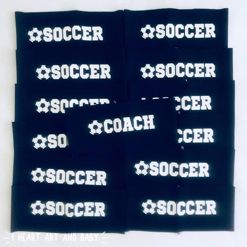 以下が含まれることがあります： 白いサッカーボールと文字が書かれた黒い布製のリストバンドの山。リストバンドの文字は「SOCCER」と「COACH」です。