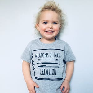 Puede incluir: Una camiseta gris con un gr&aacute;fico negro de un cuaderno con el texto "Weapons of Mass Creation" y un dibujo de l&aacute;pices, bol&iacute;grafos y pinceles.