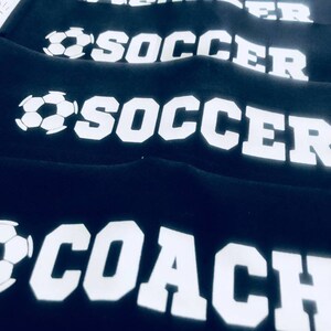 以下が含まれることがあります： 白いサッカーボールと文字が書かれた黒い布製のリストバンドが4つ。 上の2つのリストバンドには「SOCCER」と書いてあり、下の2つのリストバンドには「SOCCER」と「COACH」と書いてあります。