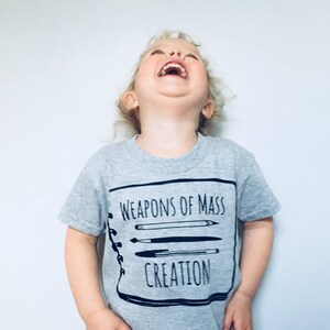 Puede incluir: Un ni&ntilde;o peque&ntilde;o con una camiseta gris con un gr&aacute;fico negro que dice "Weapons of Mass Creation" y un dibujo de un cuaderno con l&aacute;pices y bol&iacute;grafos dentro.