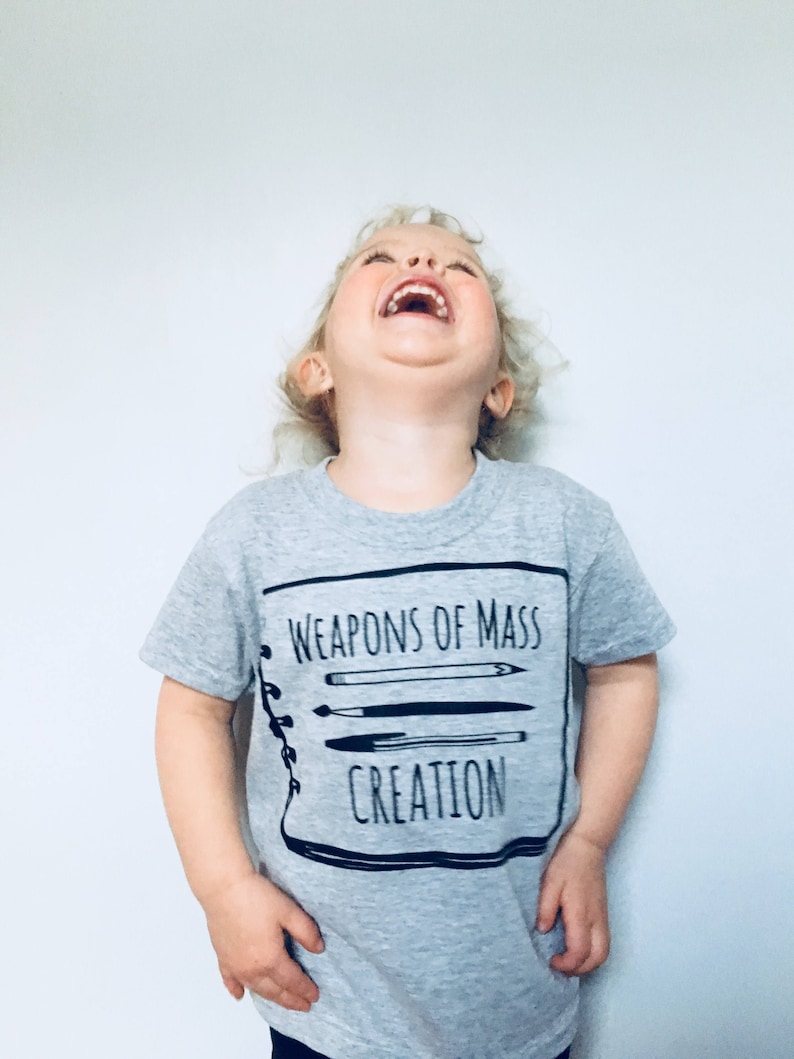 Puede incluir: Un ni&ntilde;o peque&ntilde;o con una camiseta gris con un gr&aacute;fico negro que dice "Weapons of Mass Creation" y un dibujo de un cuaderno con l&aacute;pices y bol&iacute;grafos dentro.
