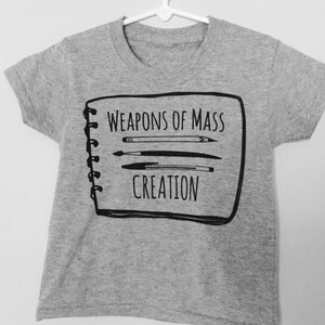Puede incluir: Camiseta gris con un gr&aacute;fico negro de un cuaderno con el texto "Weapons of Mass Creation" y un dibujo de pinceles.