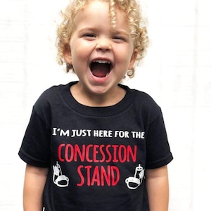 以下が含まれることがあります： 黒いTシャツを着た子供。Tシャツには「I'm just here for the concession stand」という文字が白と赤で書かれており、ホットドッグと飲み物のグラフィックが描かれている。