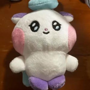May include: A plush toy in the shape of a cartoon character. The toy is primarily white with pink cheeks, purple feet and ears, and a light blue halo. It has large black eyes and a small smiling mouth.