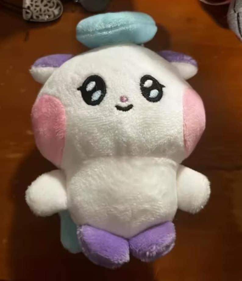 May include: A plush toy in the shape of a cartoon character. The toy is primarily white with pink cheeks, purple feet and ears, and a light blue halo. It has large black eyes and a small smiling mouth.