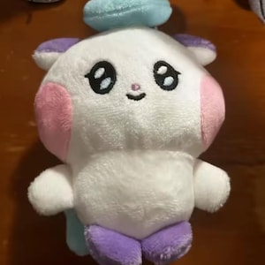 May include: A plush toy in the shape of a cartoon character. The toy is primarily white with pink cheeks, purple ears and feet, and a light blue halo. It has large black eyes and a small smiling mouth.