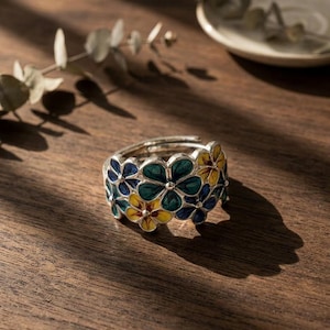 Könnte beinhalten: Ein silberner Ring mit floralem Design. Der Ring zeigt eine Reihe von Blumen in Blau-, Grün- und Gelbtönen. Der Ring liegt auf einer Holzoberfläche, mit einem unscharfen Hintergrund.