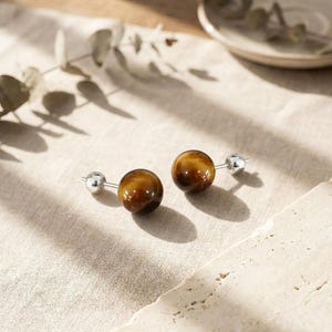 Pode incluir: Um par de brincos de tarracha com pedras preciosas olho-de-tigre esféricas e polidas em tons de castanho dourado e castanho escuro, montadas em hastes de metal prateado com fechos esféricos pequenos a condizer.